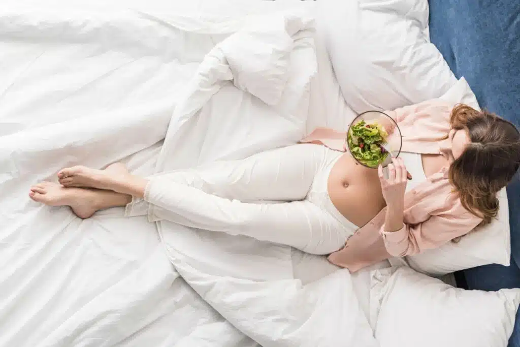 Kobieta w ciąży je sałatkę leżąc na łóżku 